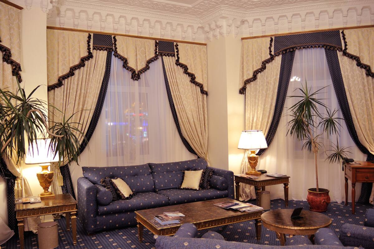 A room with a blue patterned sofa, a wooden coffee table, and two lamps. There are curtains with a decorative top and a view of the city through the windows.
