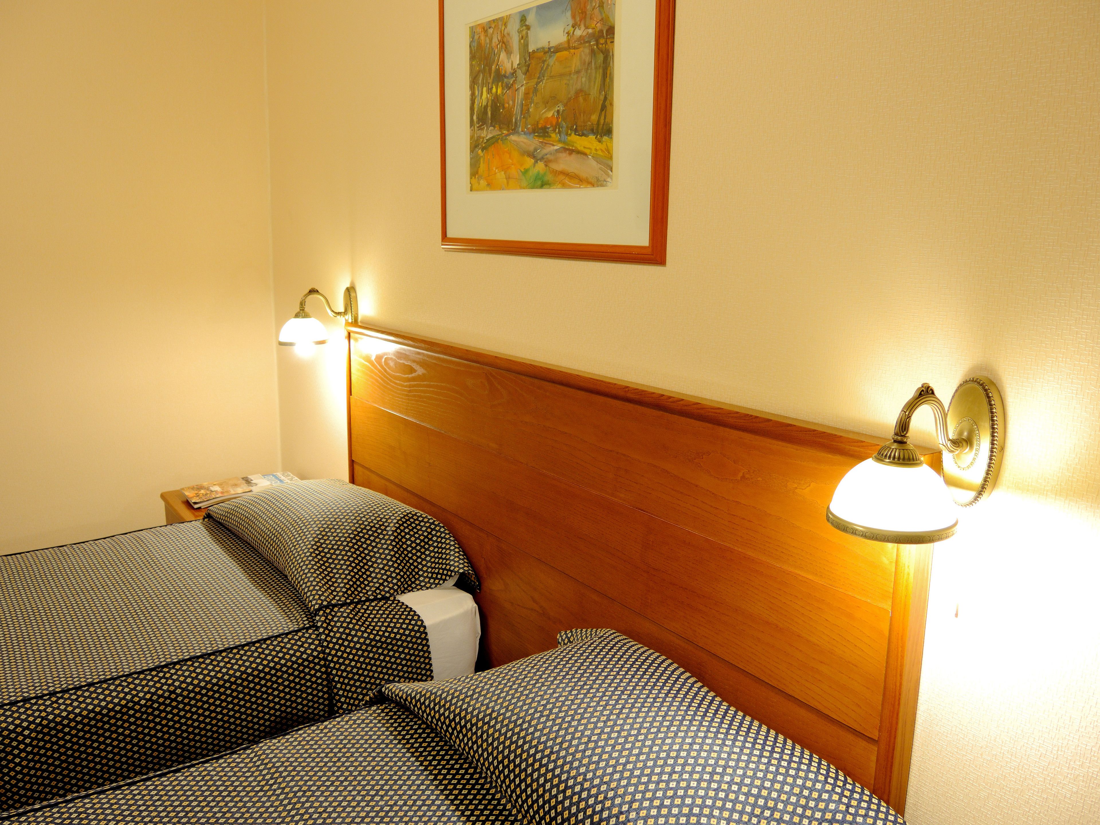 Habitación de hotel con dos camas individuales, cabeceros de madera y lámparas de lectura fijadas a la pared.