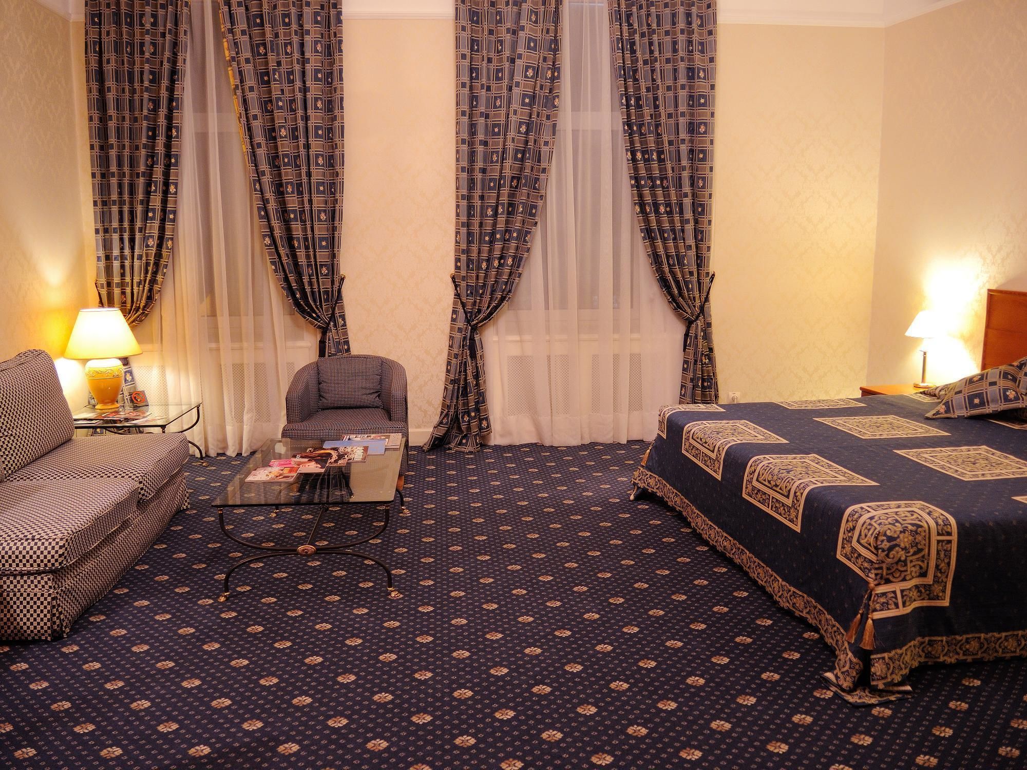 Una habitación de hotel con alfombra estampada, un sofá, una silla, una mesita con una lámpara y una cama con edredón estampado.