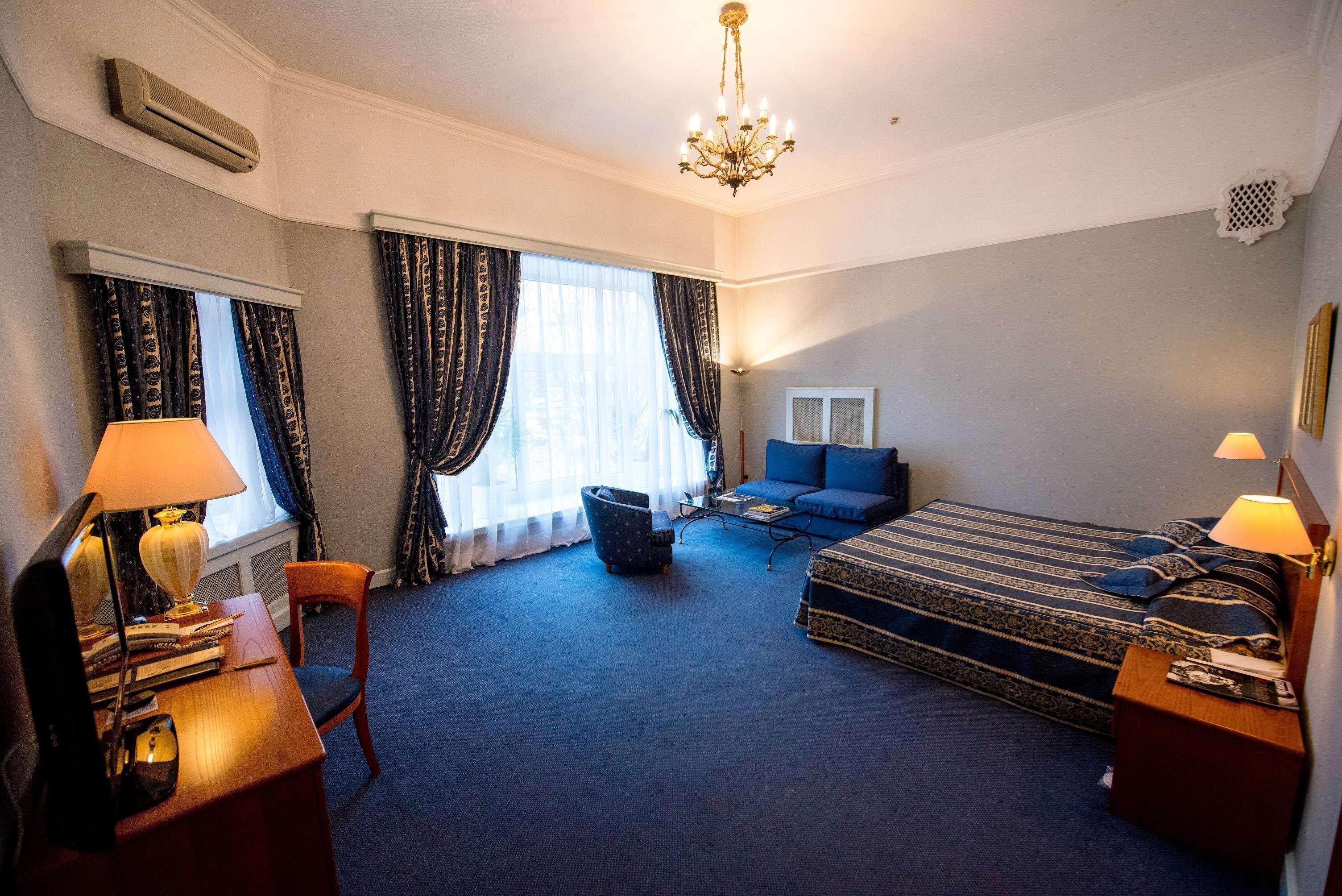 A hotel room with a bed, a desk with a lamp, a chair, a sofa, and a chandelier.