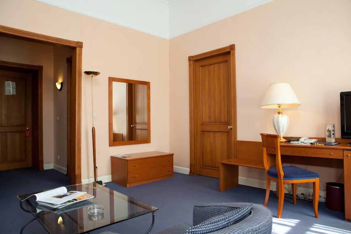 A hotel room with a wooden desk, a chair, a glass coffee table, a lamp, a mirror, and a wardrobe.