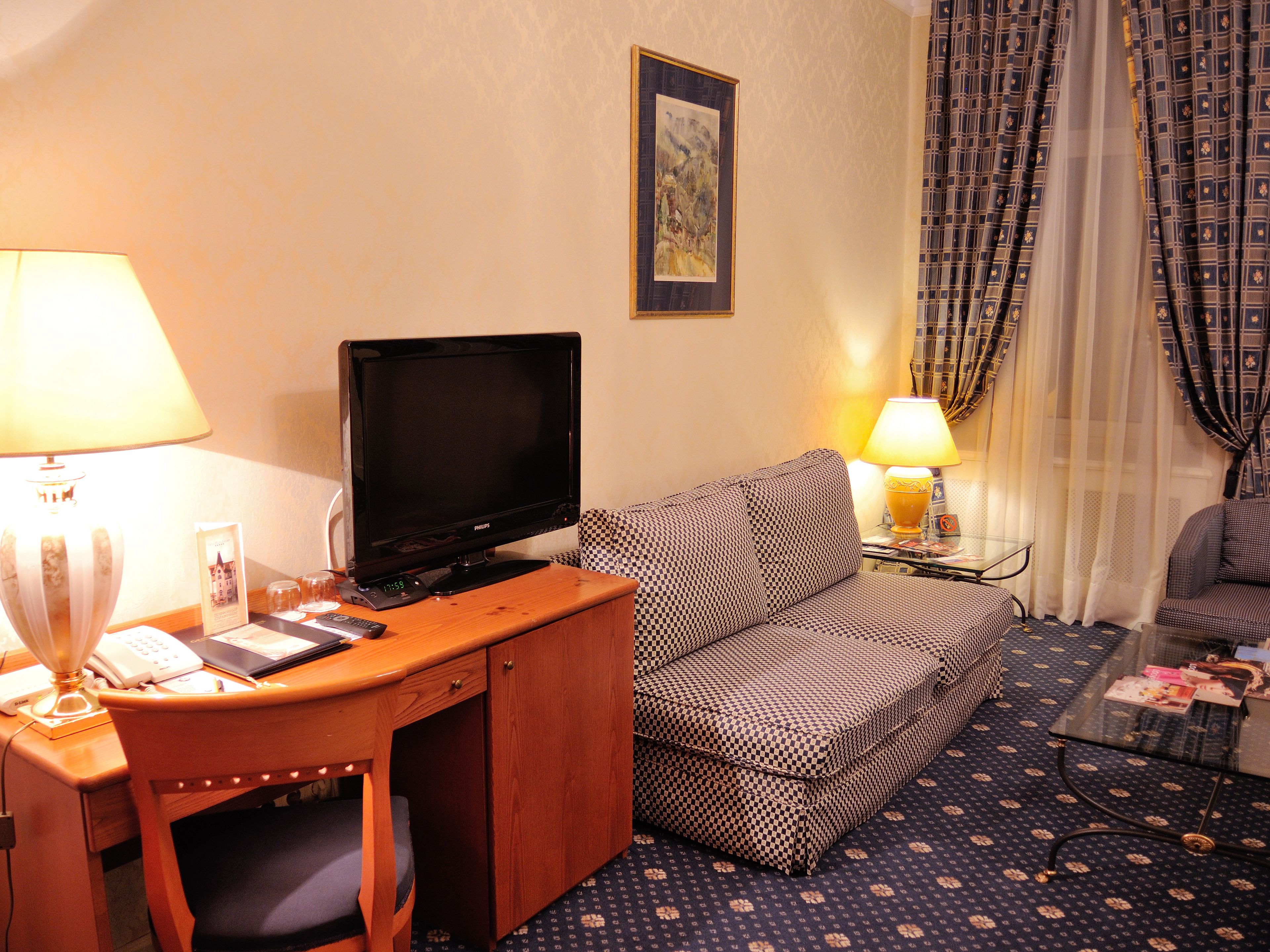 Ein Hotelzimmer mit einem Holztisch, einem Fernseher, einem gemusterten Sofa, einem kleinen Tisch mit Lampe und einem Glastisch.