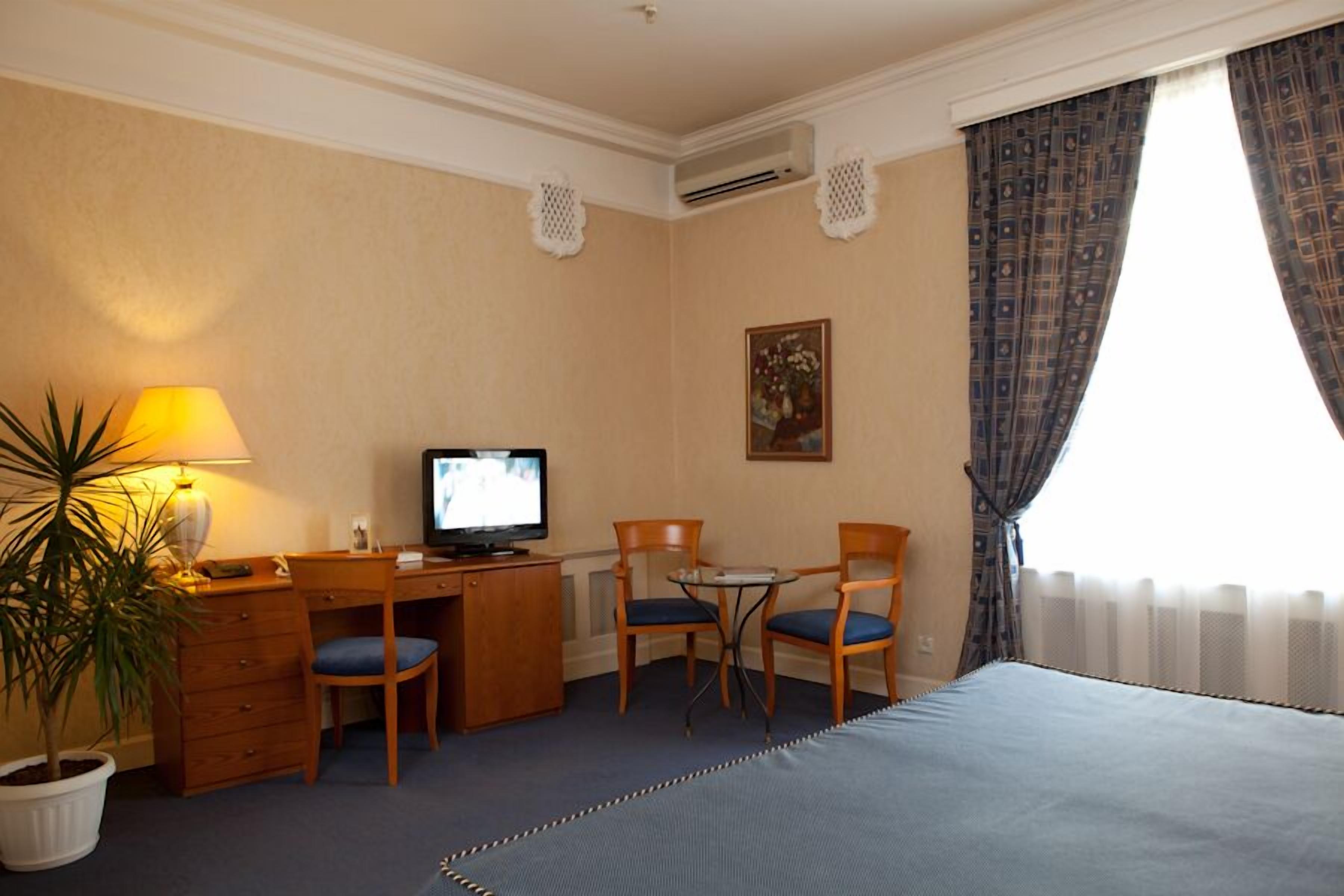 A hotel room with a bed, desk, two chairs, a television, a potted plant, and a painting on the wall.