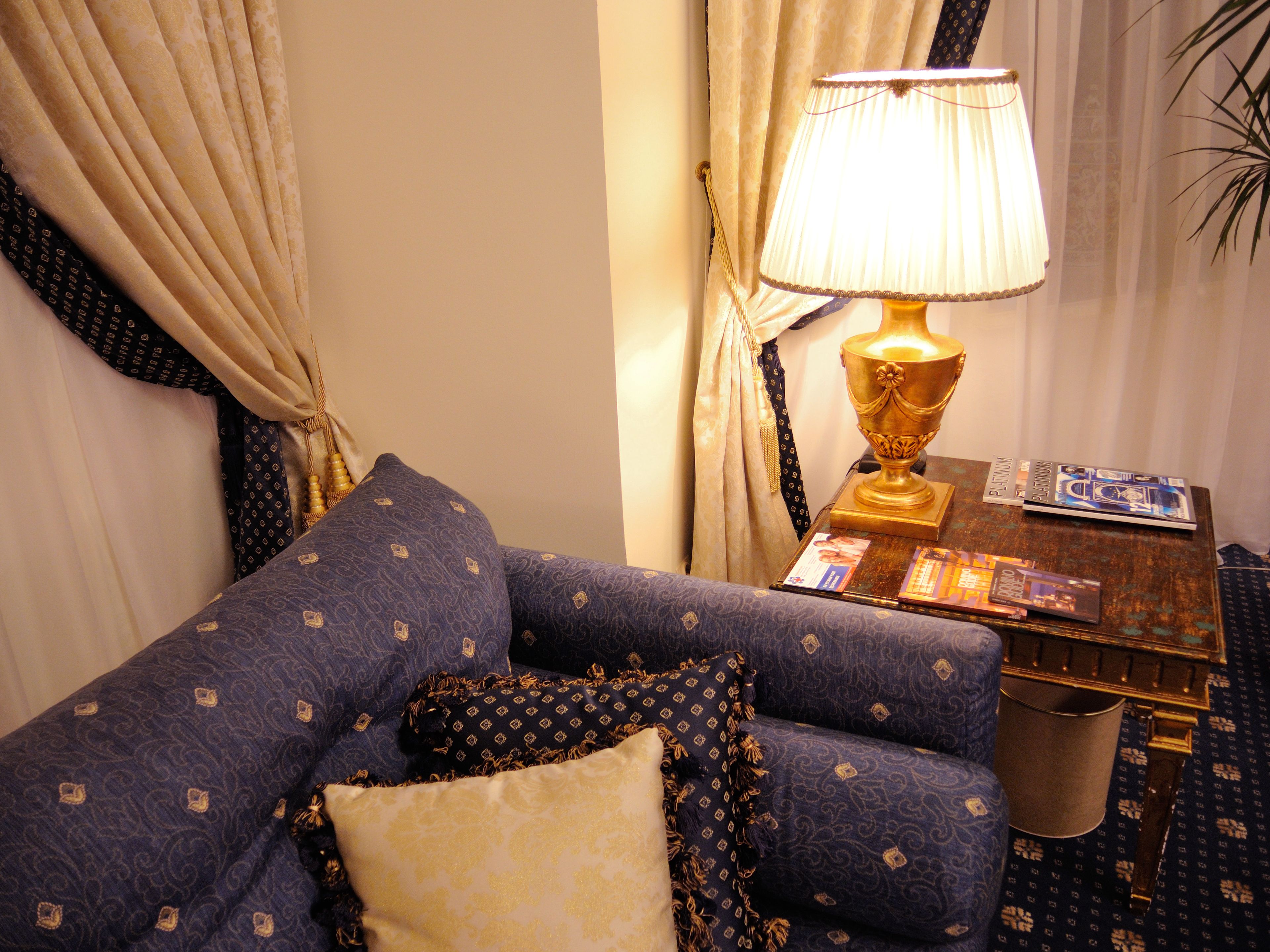 A room with a blue patterned sofa, a gold lamp, and a small table with books and a lamp.