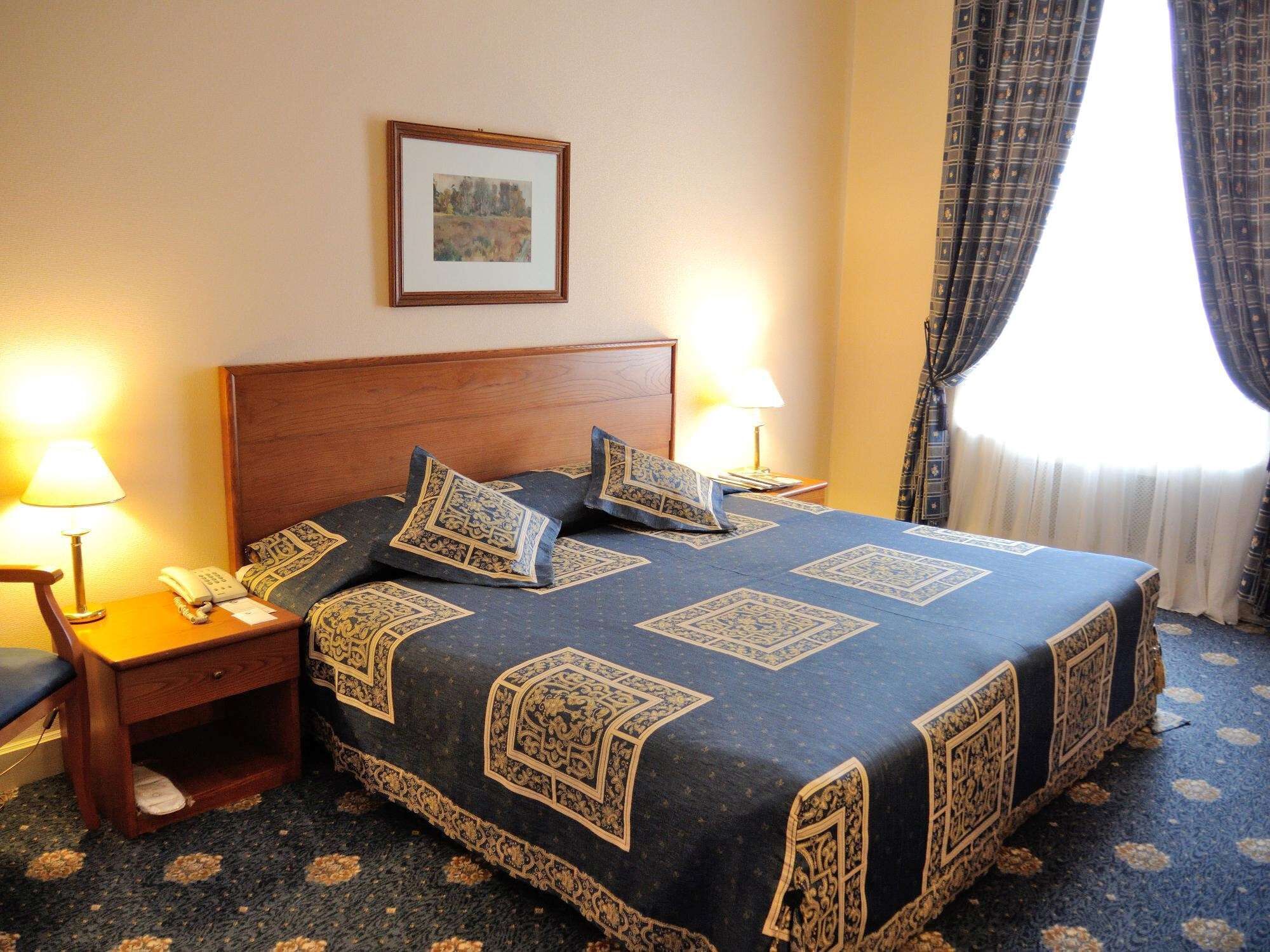 A hotel room with a large bed, two bedside tables with lamps, a nightstand, a framed picture on the wall, and a window with curtains.