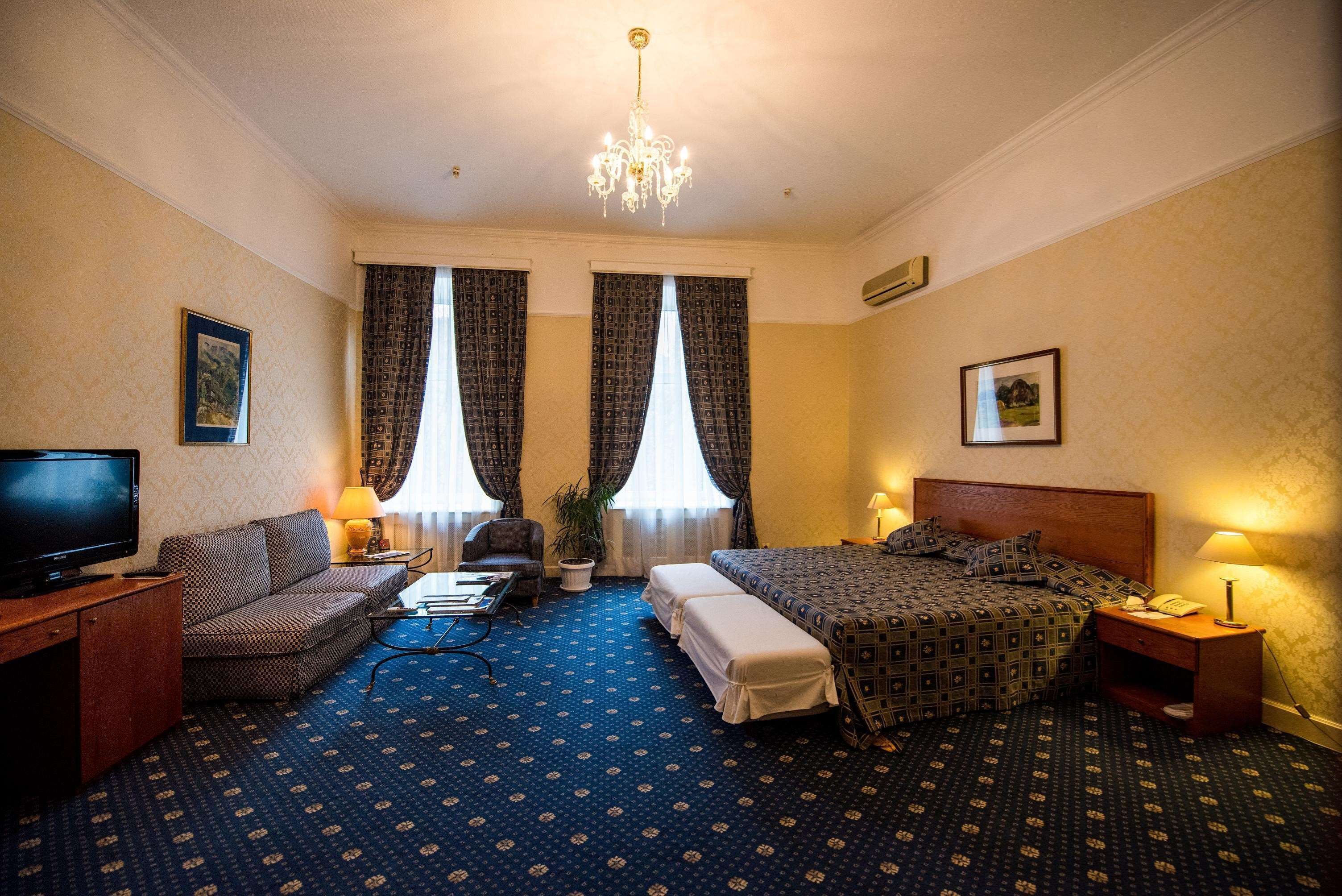 A hotel room with a large bed, a sofa, a desk with a TV, and a chandelier.