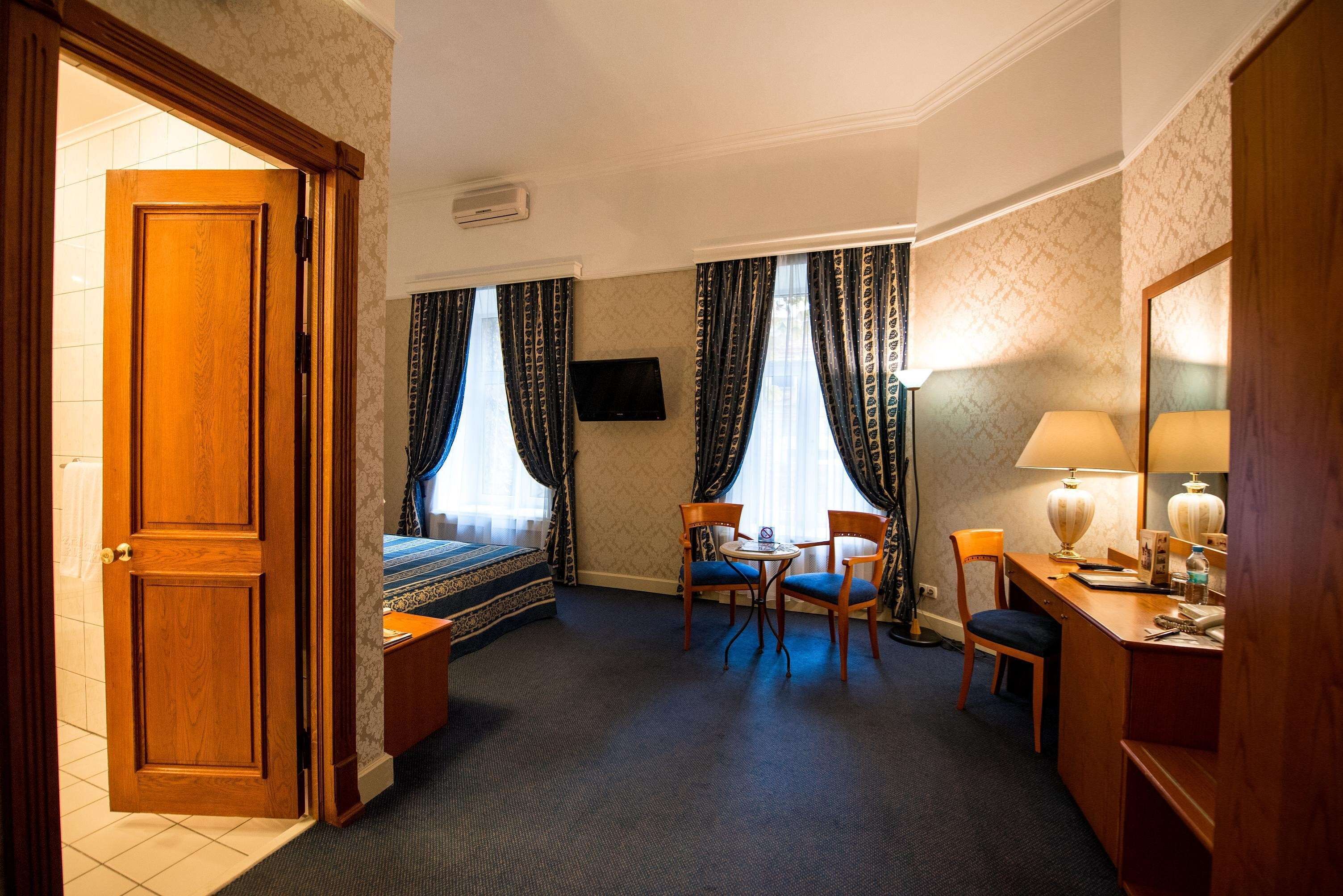 A hotel room with a bed, a desk with a chair, a small table, a lamp, and a television.