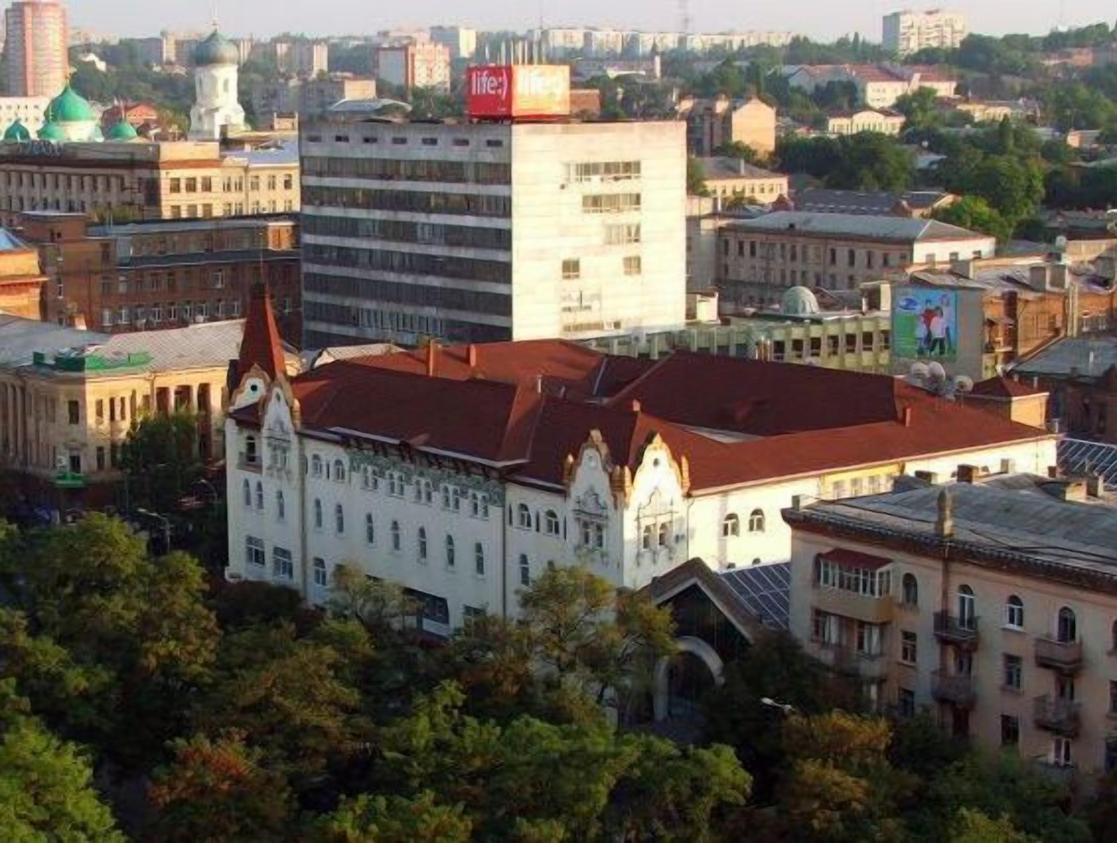 Un paisaje urbano con un edificio blanco destacado, que cuenta con una torre de reloj y un techo rojo, rodeado de otros edificios y vegetación.