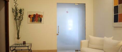 A living room with a white sofa, a glass coffee table, a framed floral artwork, and a door with a 'no entry' sign.