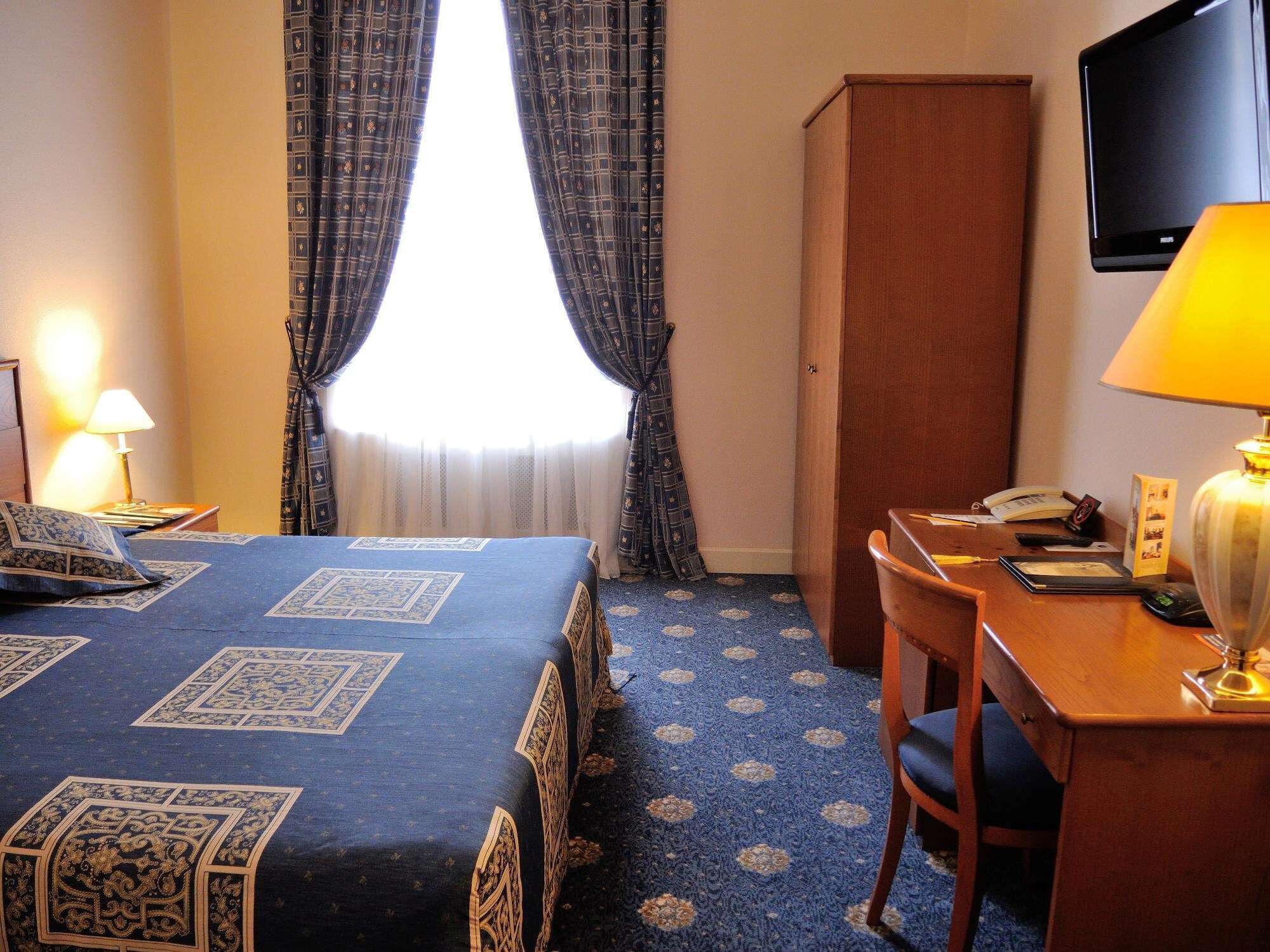 Ein Hotelzimmer mit Bett, Schreibtisch, Stuhl, Kleiderschrank, Fernseher und einem Fenster mit Vorhängen.