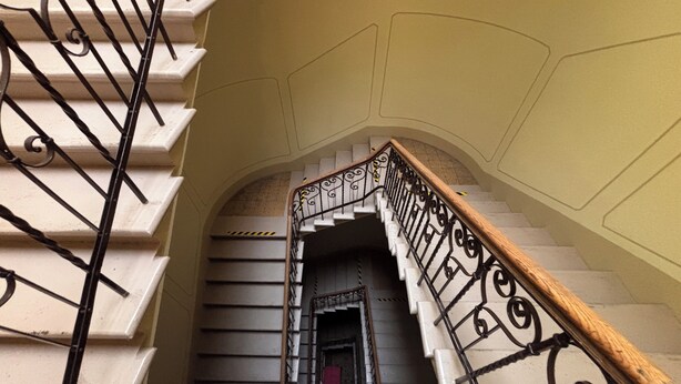 Una escalera con barandales de metal negro ornamentados y barandilla de madera, que conduce a un arco de entrada.