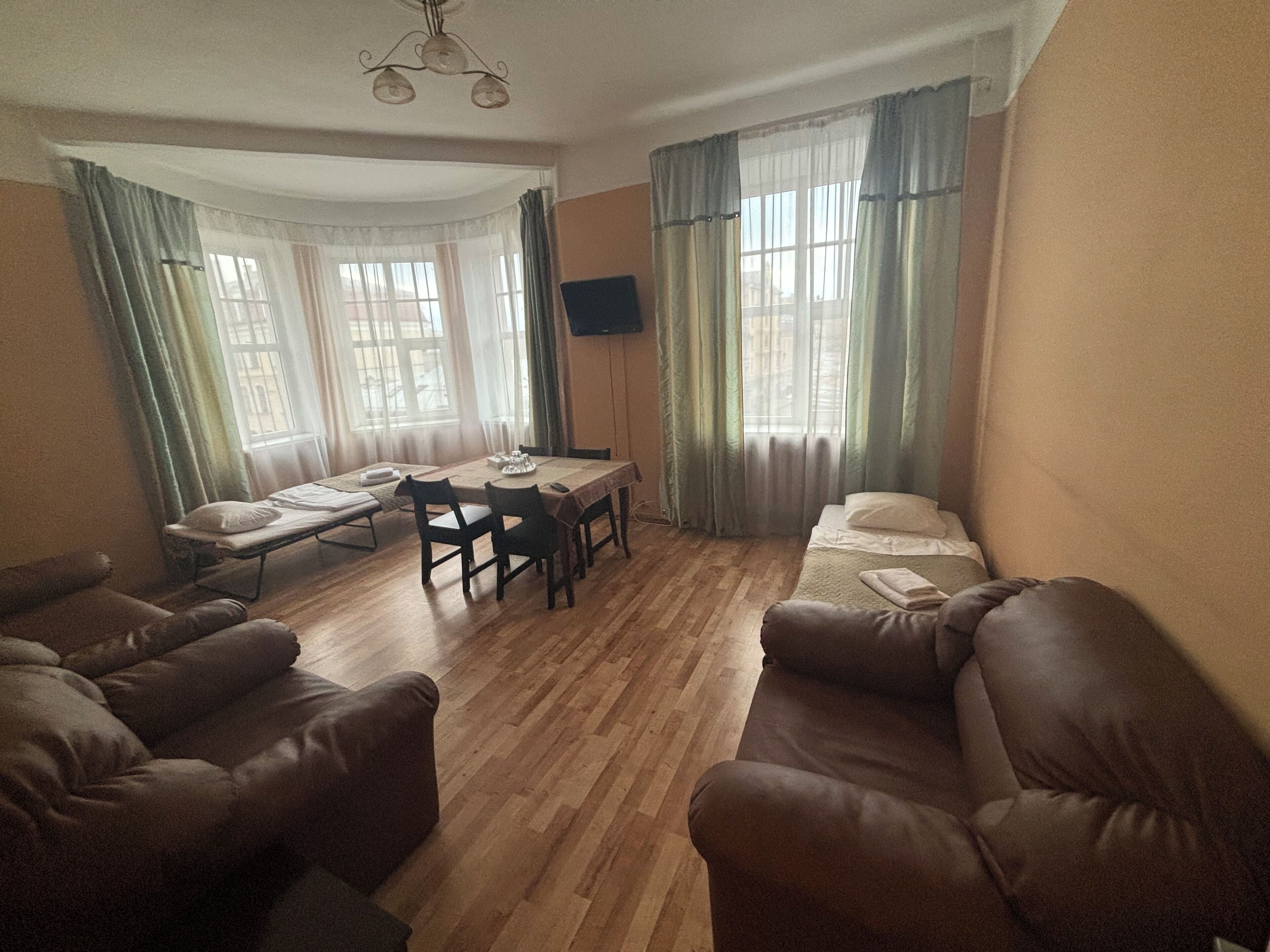 A room with a wooden floor, a dining table with chairs, a sofa, a bed, and a TV mounted on the wall.