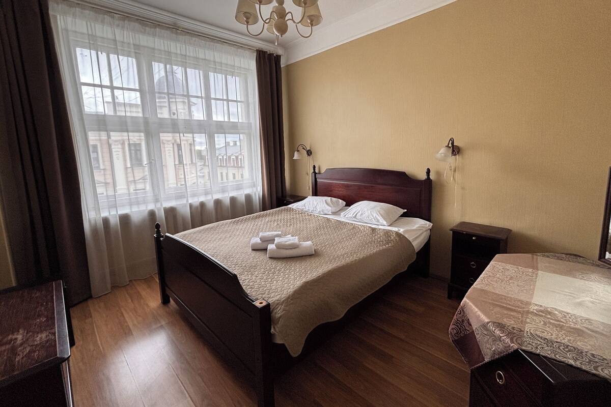 A hotel room with a large bed, a bedside table, a nightstand, a dresser, a window with curtains, and a chandelier.