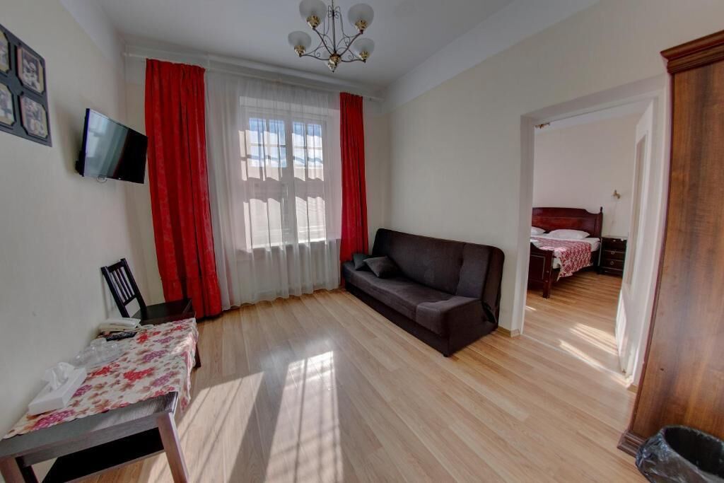 Een kamer met een bank, een kleine tafel met een bloemrijk tafelkleed, een aan de muur bevestigde televisie, en een raam met rode gordijnen.