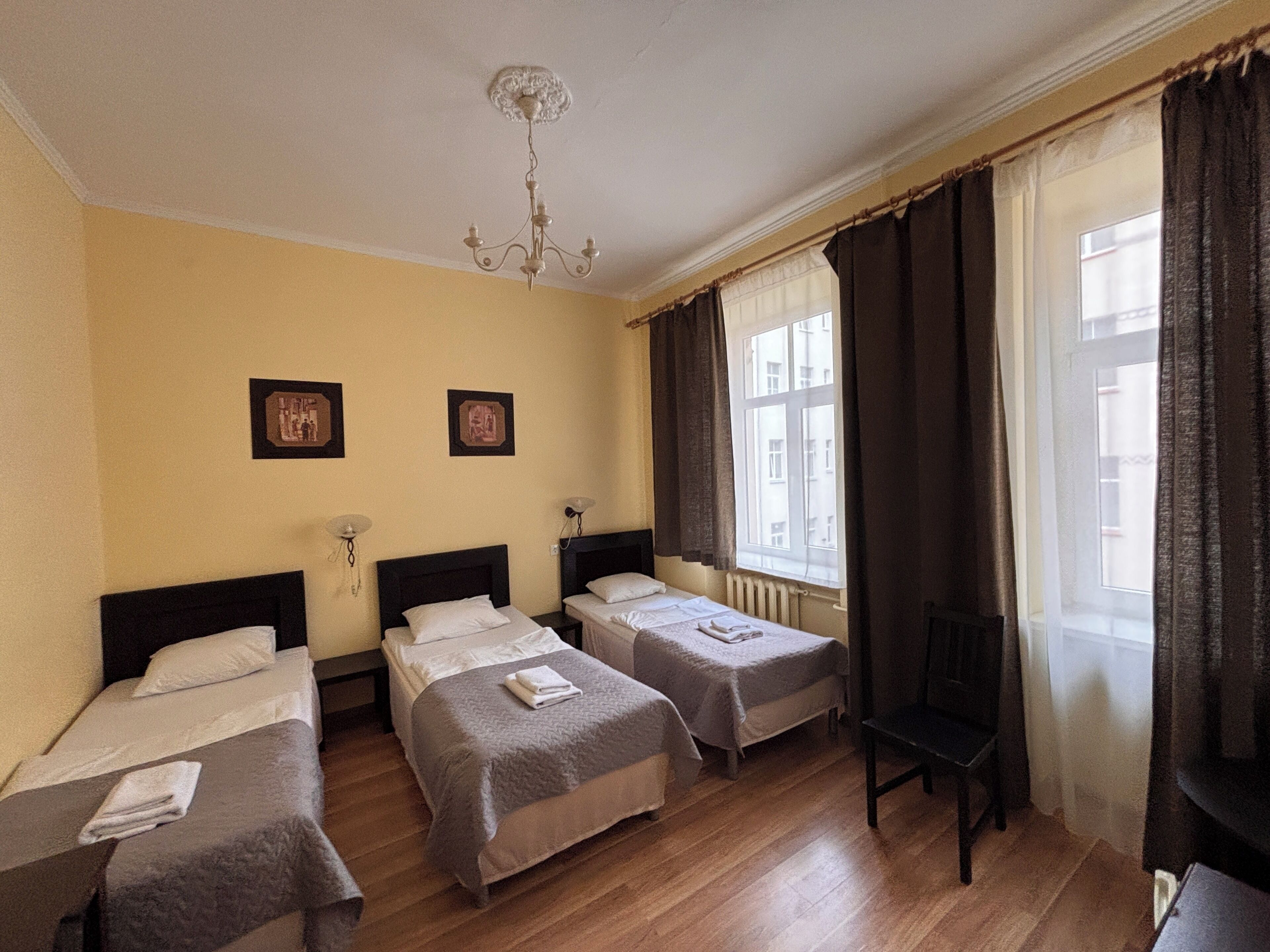 Une chambre d’hôtel avec trois lits simples, un plancher en bois, un lustre et une fenêtre avec des rideaux.