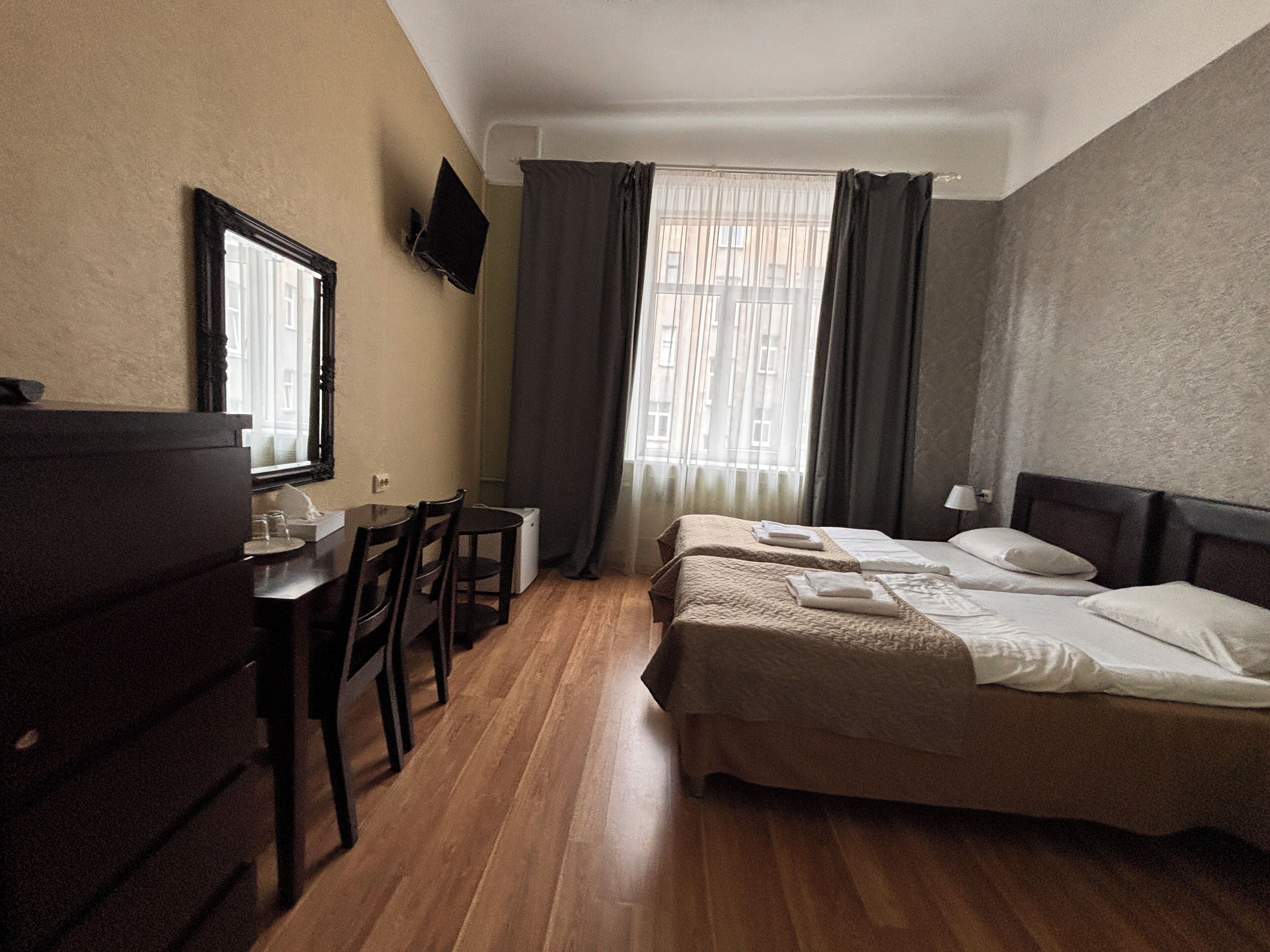 A hotel room with a wooden floor, a bed with white linens, a desk with a chair, a television mounted on the wall, and a window with curtains.