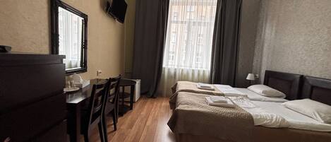 A hotel room with a wooden floor, a bed with white linens, a desk with a chair, a television mounted on the wall, and a window with curtains.
