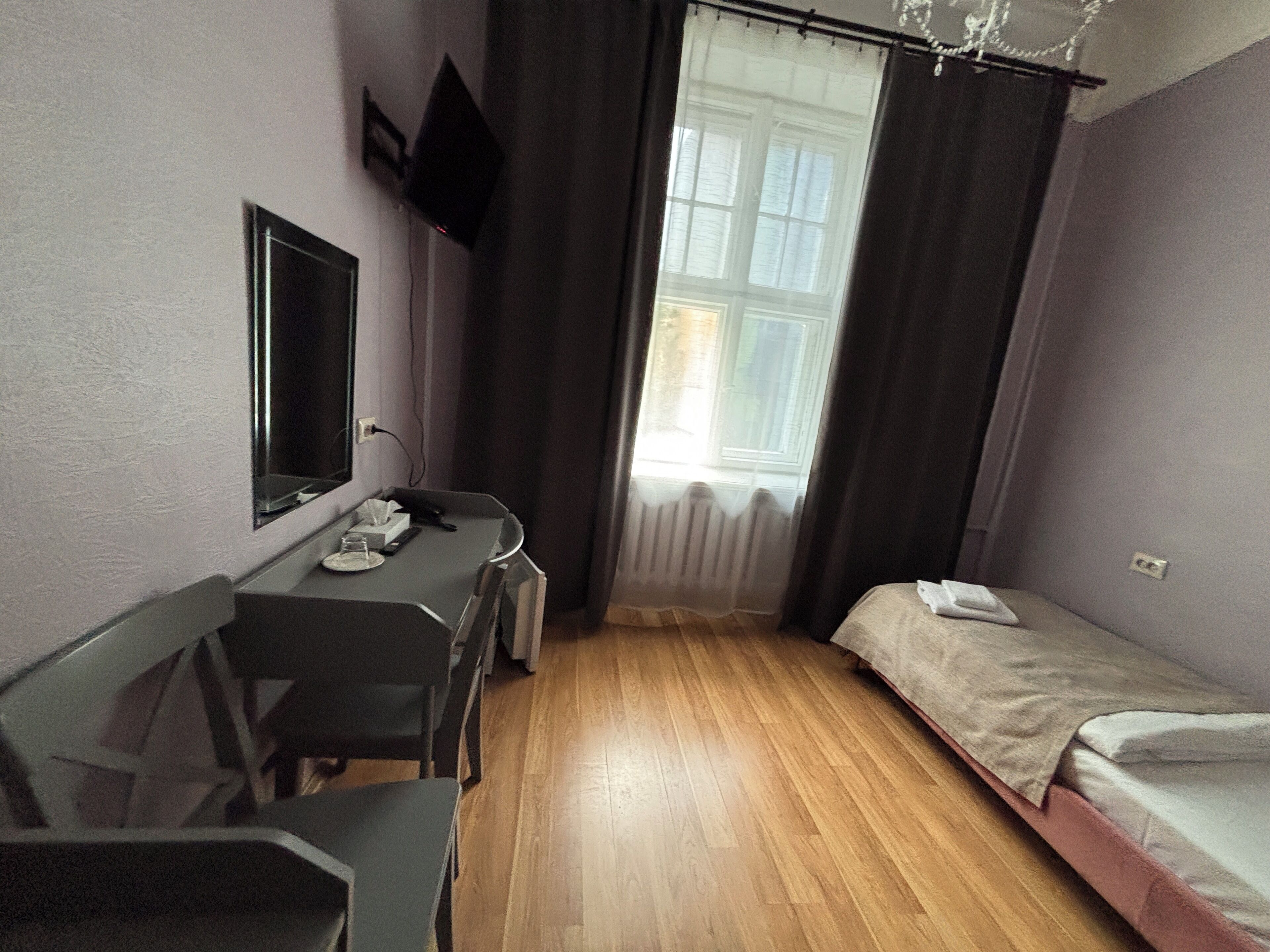 Una habitación pequeña y funcional con suelo de madera, una cama individual, un escritorio con silla, un espejo y un televisor fijado a la pared.