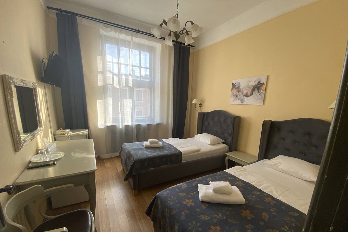A hotel room with two beds, a desk with a chair, a mirror, a window with curtains, and a chandelier.