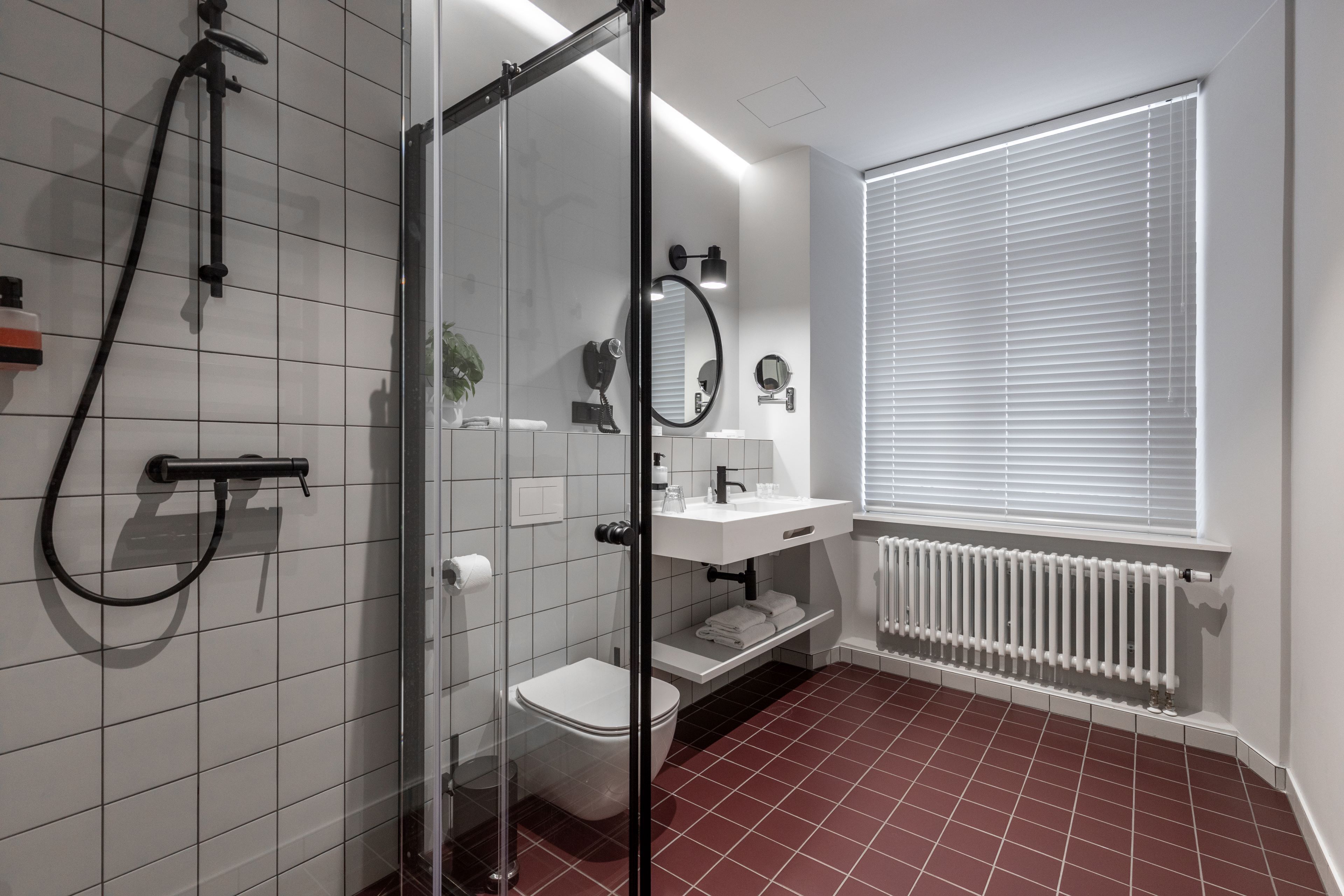 A modern bathroom with a glass shower enclosure, white tiles, a white toilet, a white sink with a mirror, and a window with blinds.