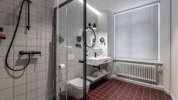 A modern bathroom with a glass shower enclosure, white tiles, a white toilet, a white sink with a mirror, and a window with blinds.