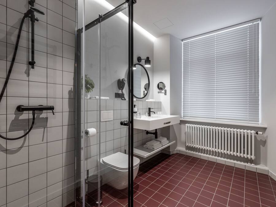 A modern bathroom with a glass shower enclosure, white tiles, a white toilet, a white sink with a mirror, and a window with blinds.
