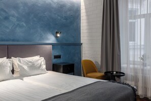A modern hotel room with a large bed, a small desk, a chair, and a window with curtains.