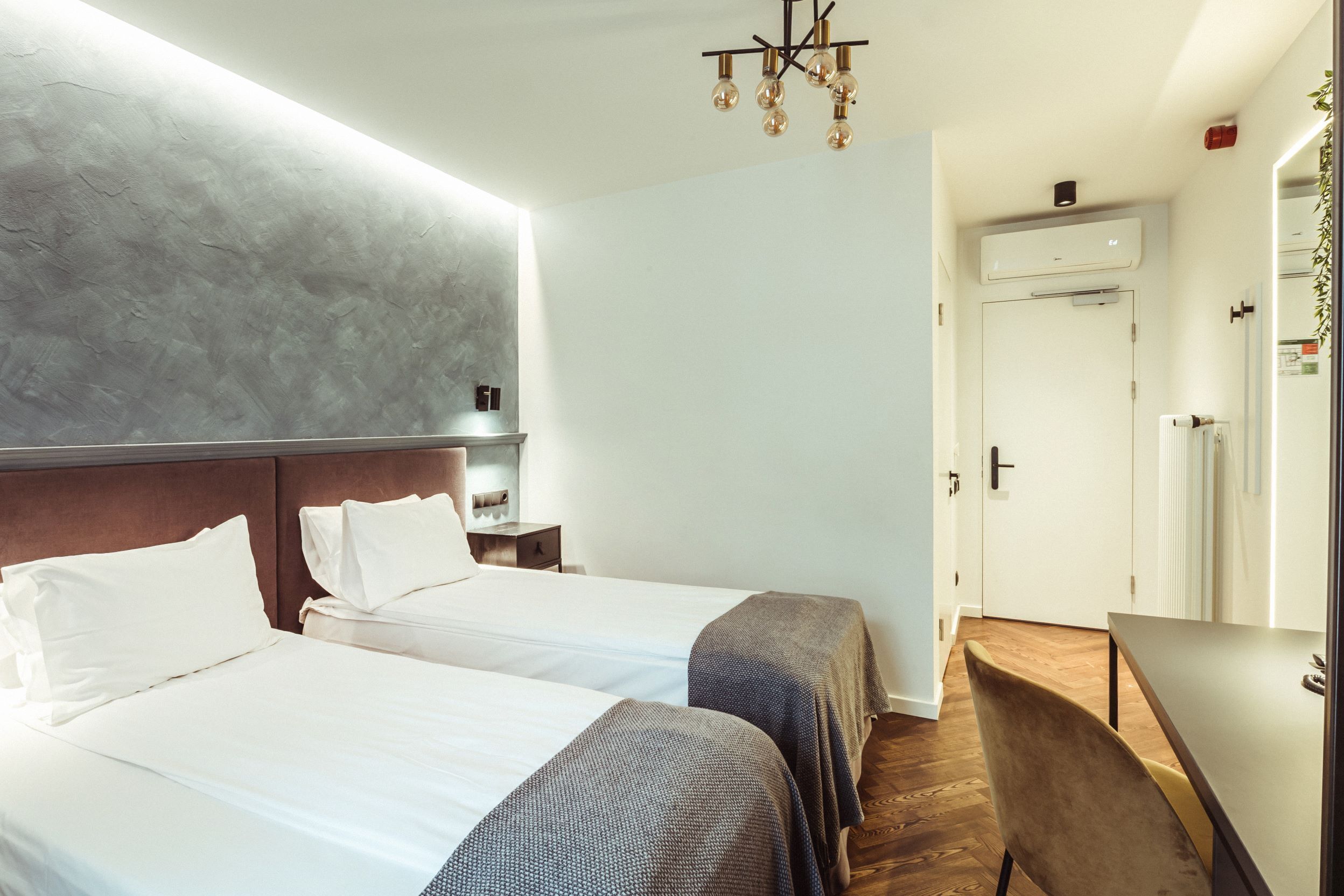 Quarto de hotel com duas camas, piso de madeira, mesa de jantar e um lustre moderno.