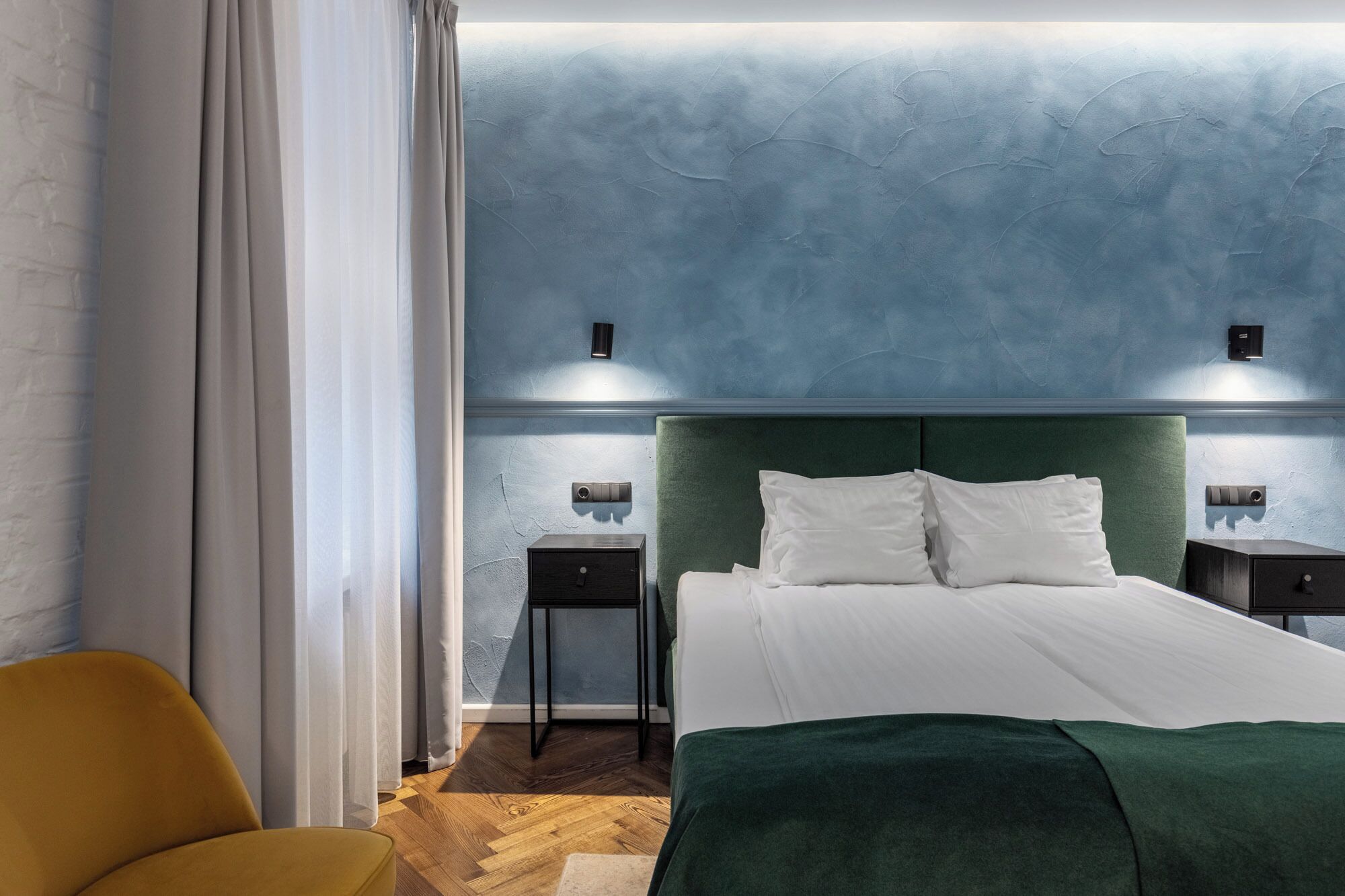 A bedroom with a green upholstered headboard, a white bedspread, two bedside tables with lamps, and a wooden floor.