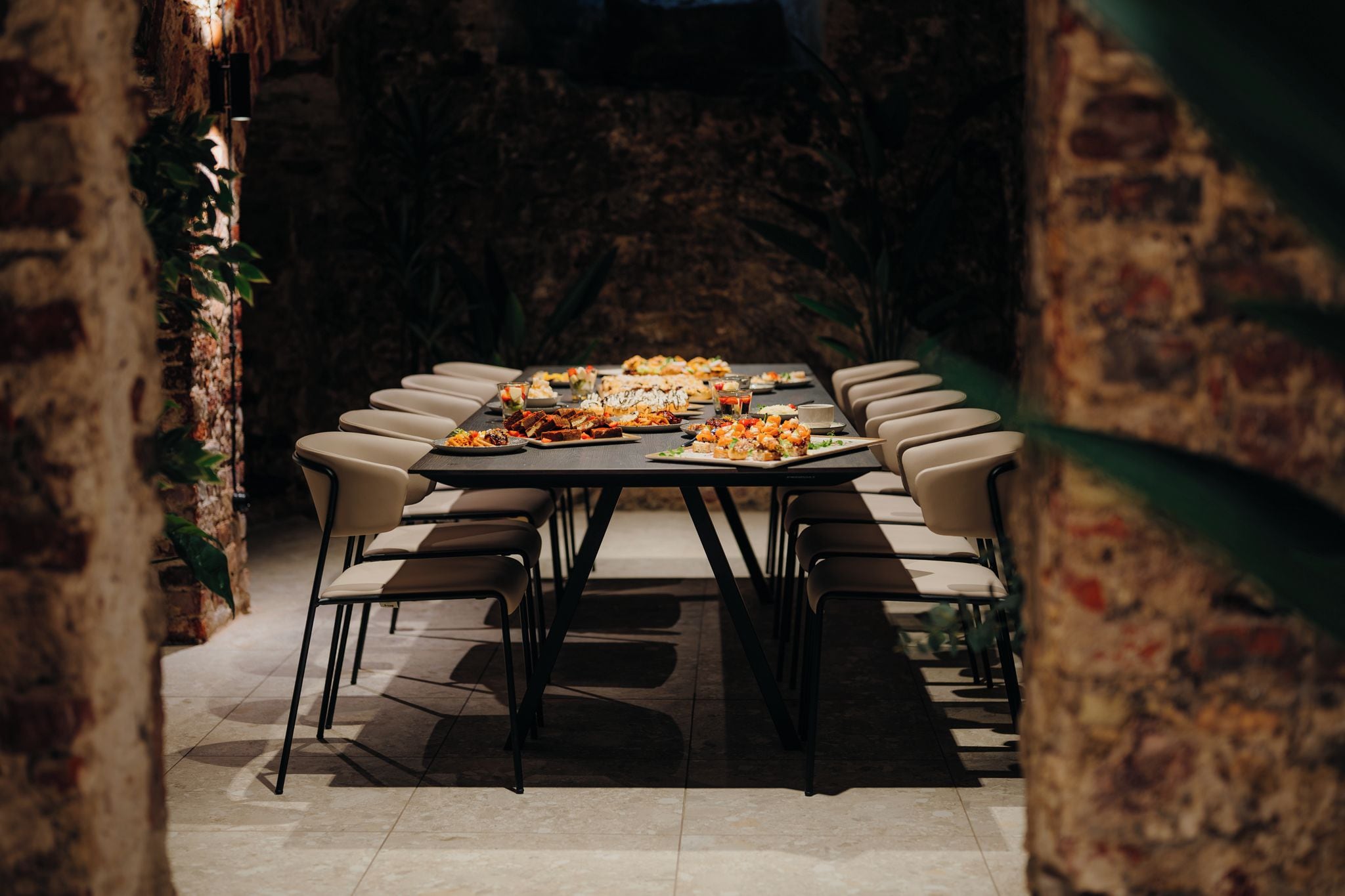 Una larga mesa de comedor con múltiples sillas, rodeada por un rústico muro de ladrillo y follaje.