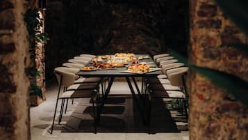 Una larga mesa de comedor con múltiples sillas, rodeada por un rústico muro de ladrillo y follaje.