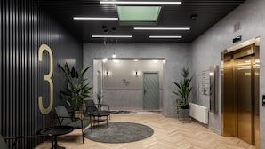 A modern reception area with a herringbone wood floor, a reception desk, two chairs, a potted plant, and an elevator.