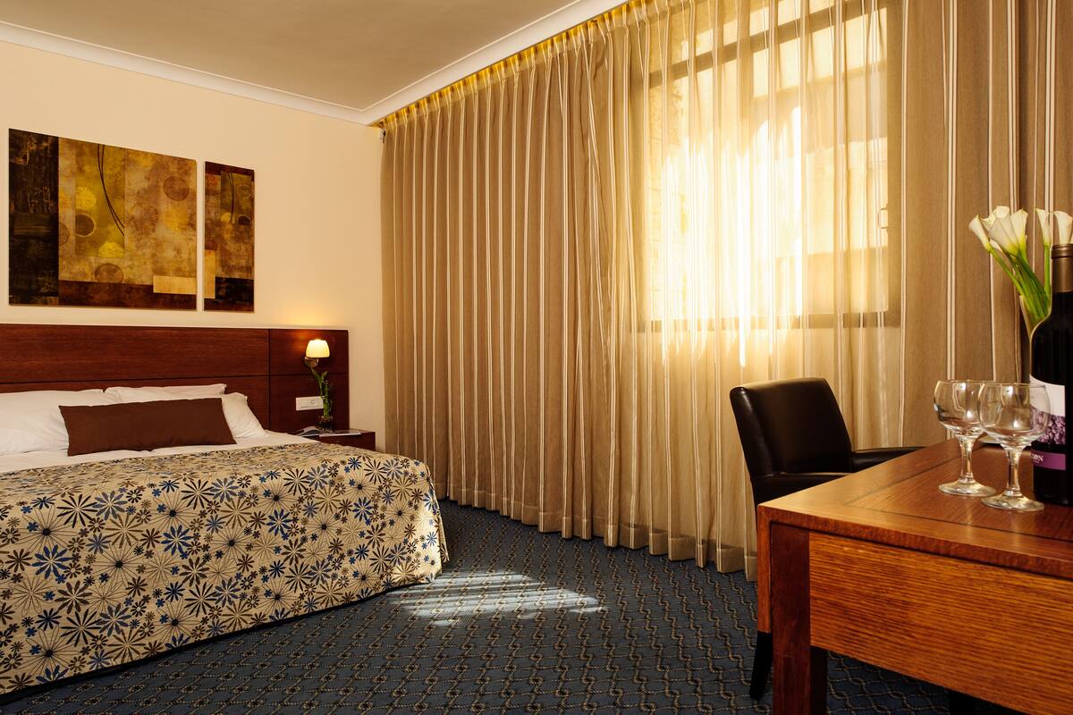 A hotel room with a bed, a desk with two glasses, a chair, and curtains.