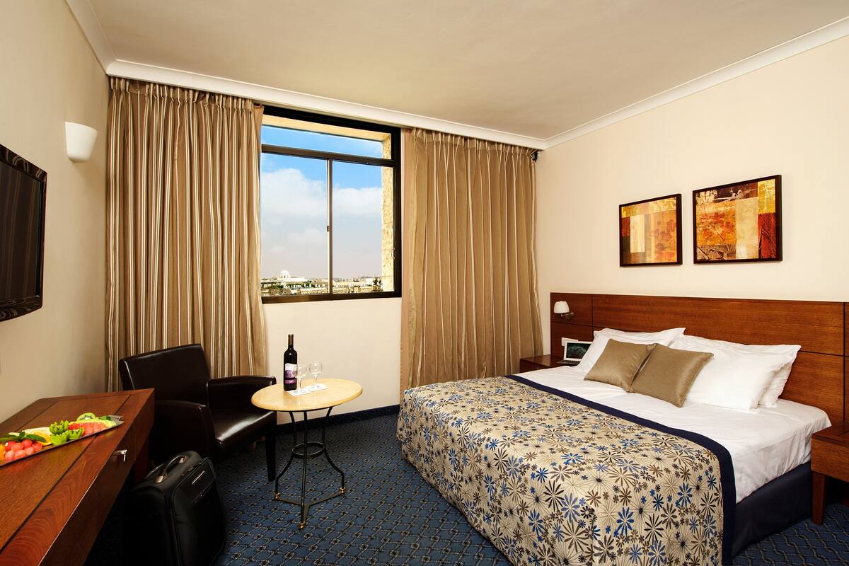 A hotel room with a bed, a desk with a chair, a small table with a bottle and glass, and two paintings on the wall.