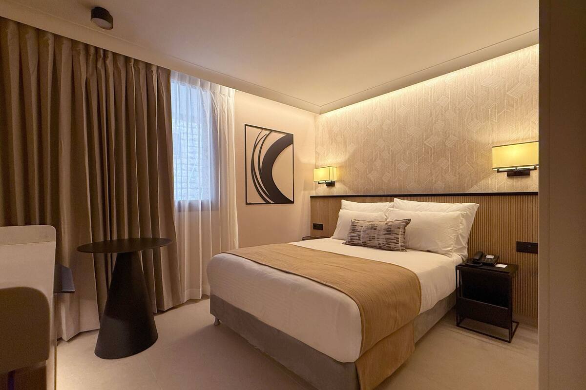 A hotel room with a bed, bedside table, lamp, and a view of the city through the window.