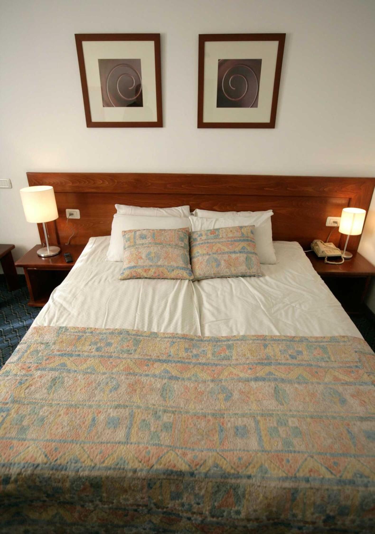 Un lit bien fait, agrémenté de coussins à motifs et d’une couverture assortie, deux tableaux encadrés au mur et des lampes de chevet de chaque côté.