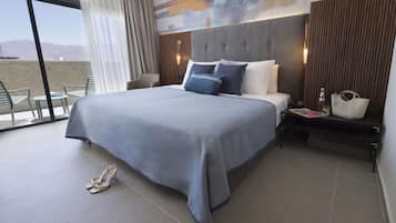 Una habitación de hotel moderna con una cama grande, vistas a las montañas, un balcón con sillas y una mesita de noche con lámpara.