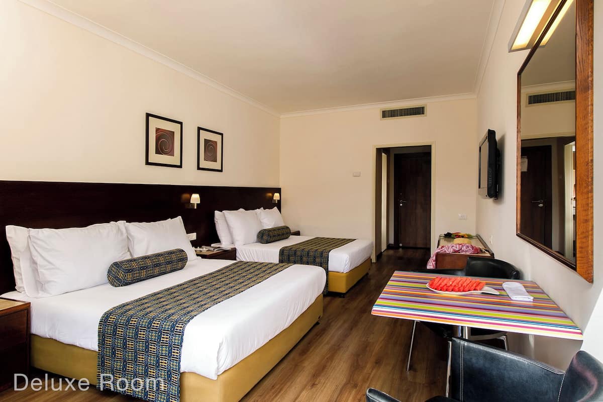 Ein Hotelzimmer mit zwei Betten, einem Schreibtisch mit gestreiftem Tischlein und einem Fernseher sowie einem Spiegel.