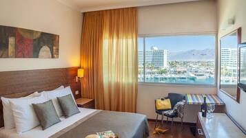 Habitación de hotel con cama, escritorio, silla, ventana grande con vistas a la ciudad y montañas, y un cuadro en la pared.
