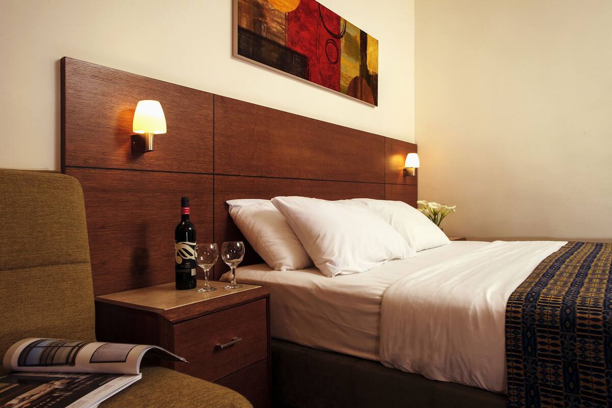 Ein Hotelzimmer mit einem hölzernen Kopfteil, einem Bett mit weißer Bettwäsche, einem Nachttisch mit einer Weinflasche und Gläsern sowie einem Gemälde über dem Bett.