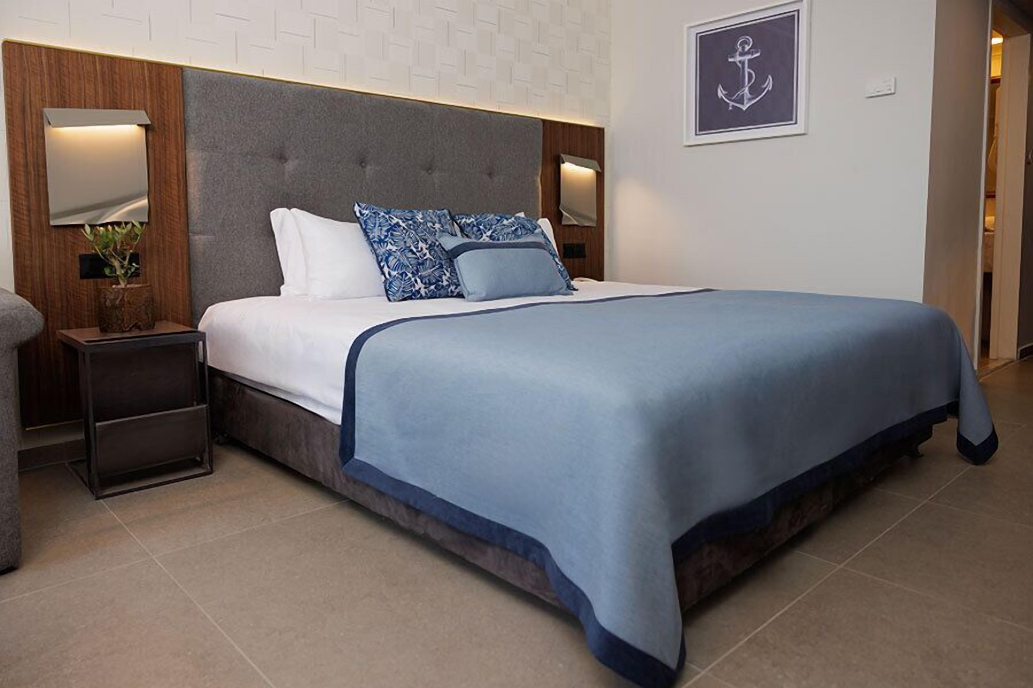 Una cama bien hecha con cabecera gris, ropa de cama blanca y detalles azules. Hay dos mesitas de noche con lámparas, un cuadro enmarcado en la pared y un suelo de baldosas.