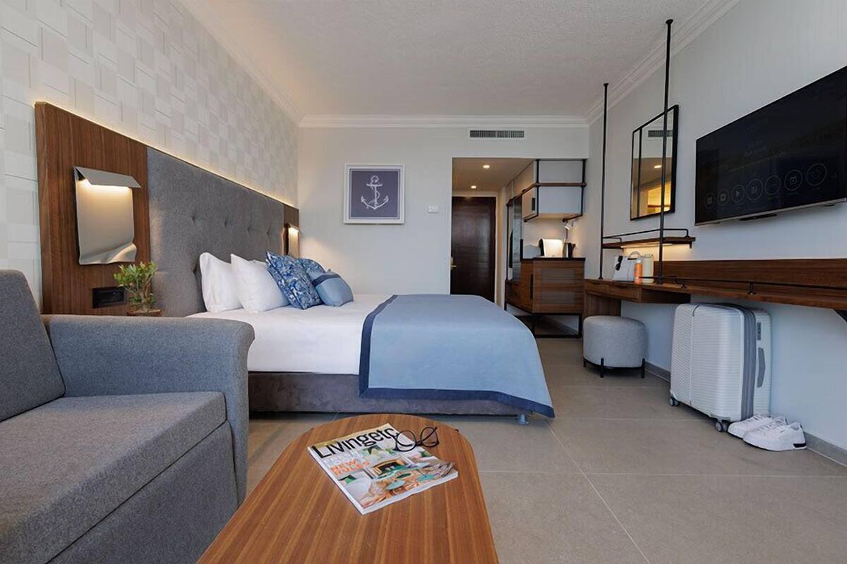 A modern hotel room with a bed, a sofa, a wooden coffee table with a magazine, a TV, and a desk with a lamp.