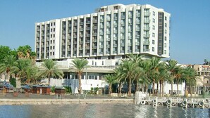 A multi-story hotel with balconies, located near a body of water with palm trees. - Caesar Premier Tiberias (Tiberias)
