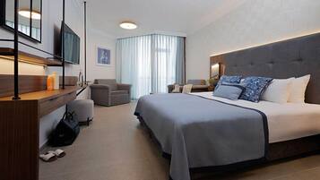 Una habitación de hotel moderna con una cama grande, un escritorio con televisor de pantalla plana, una zona de estar con dos sillones y un ventanal con cortinas.