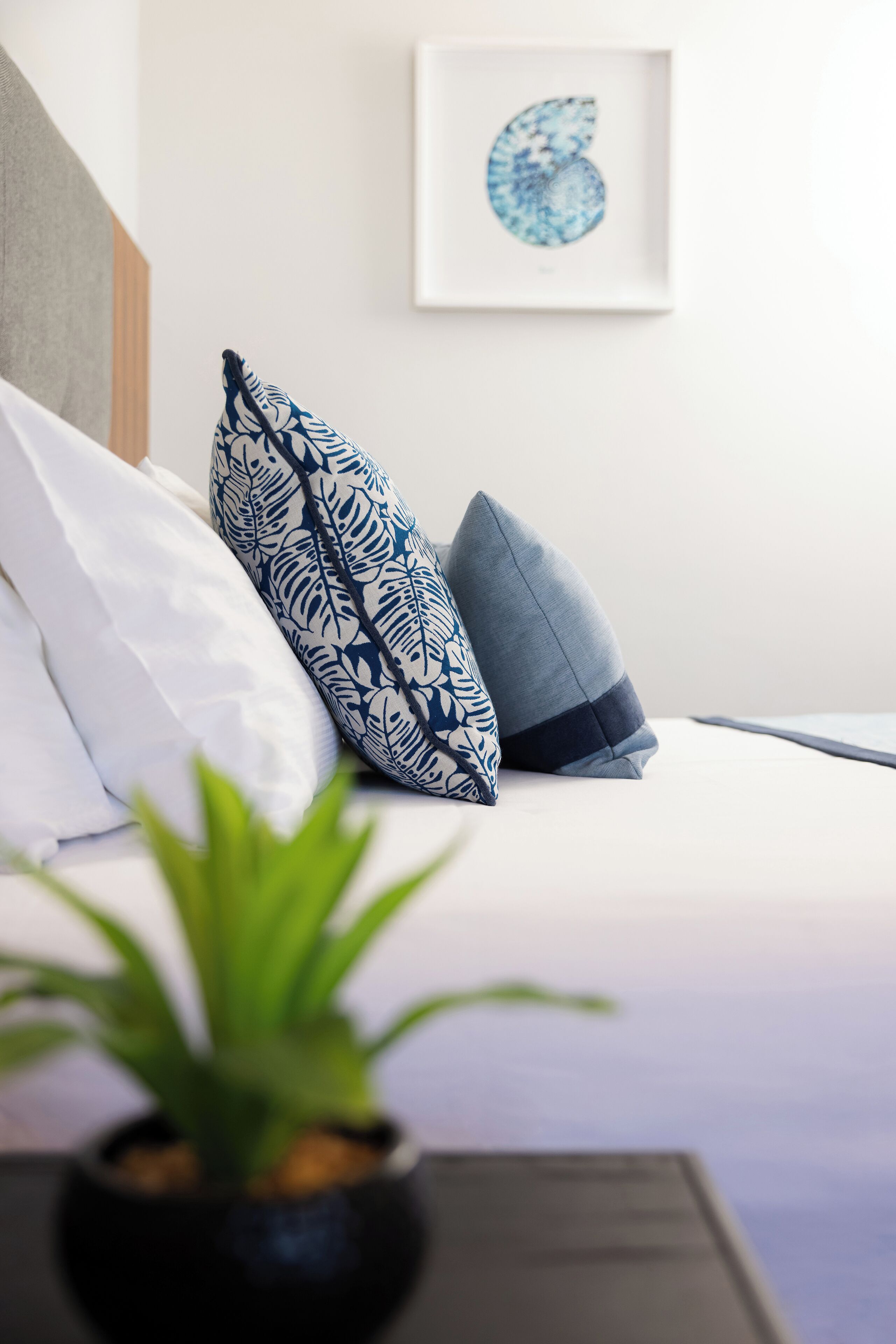 Una cama bien tendida con almohadones estampados, una obra enmarcada en la pared y una planta en maceta al frente.