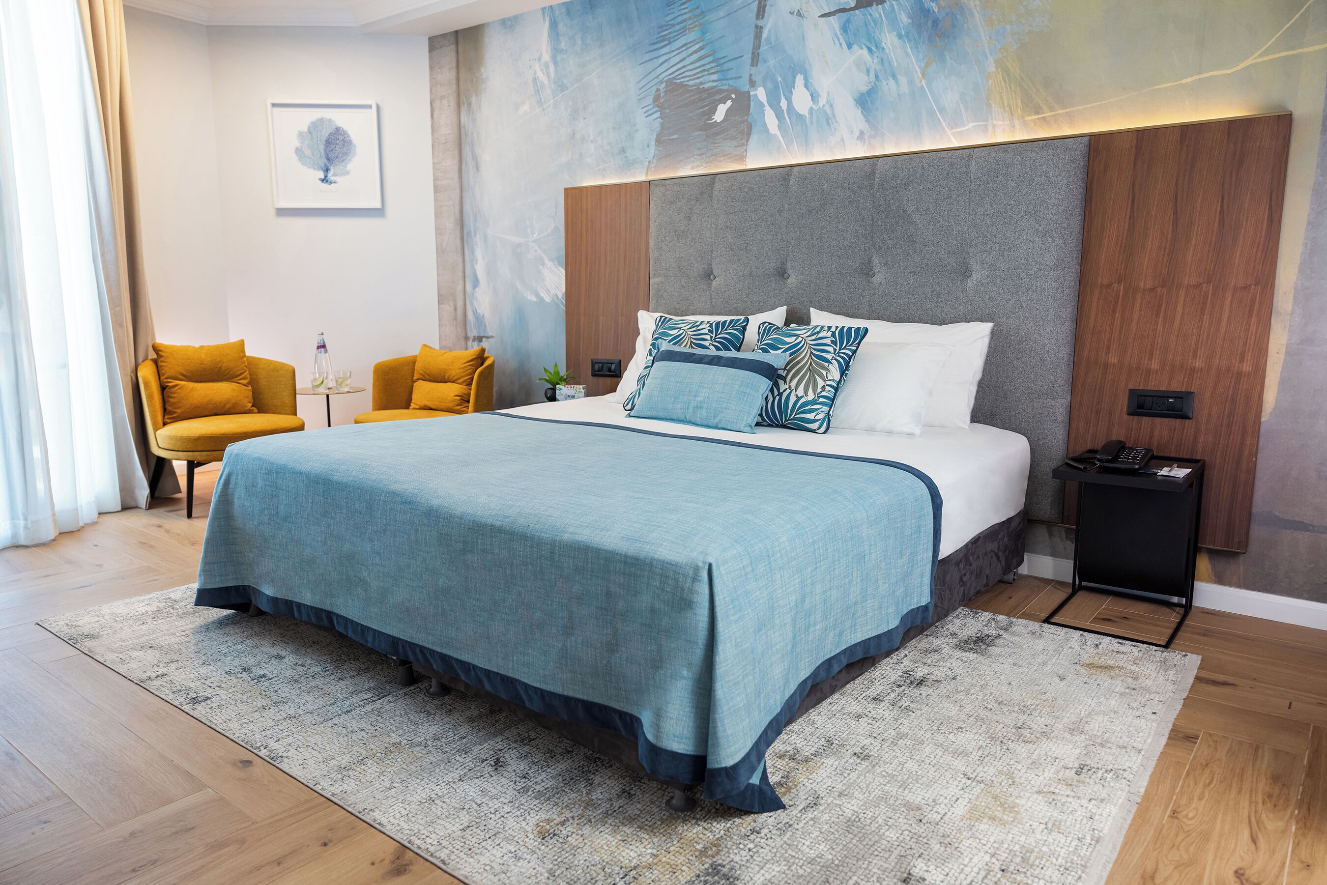 A modern bedroom with a large bed, two yellow chairs, a small table, and a wall art piece.