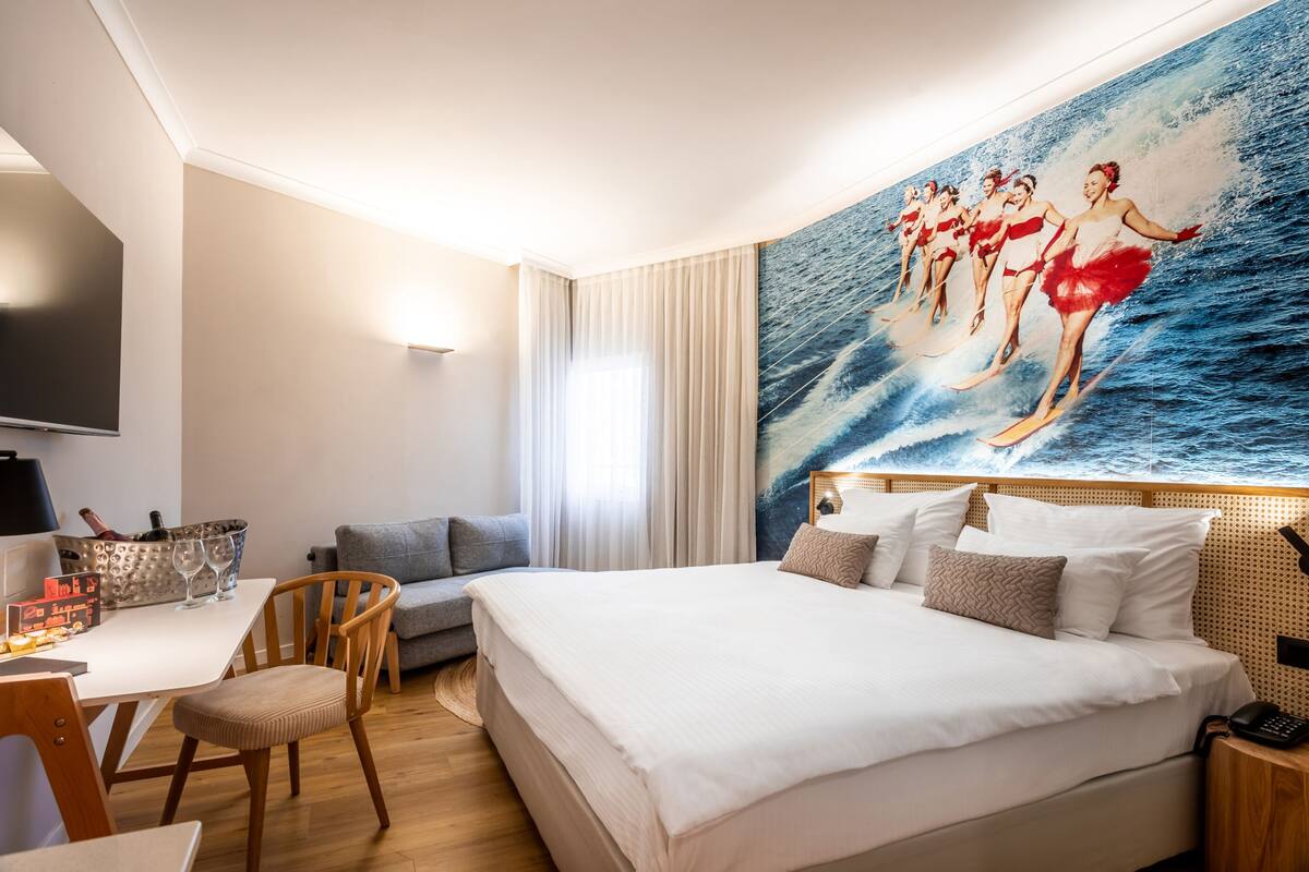 Ein Hotelzimmer mit einem großen Bett, einem Schreibtisch mit Stuhl, einem Flachbildfernseher und einem Gemälde von Tänzern auf einer Welle.