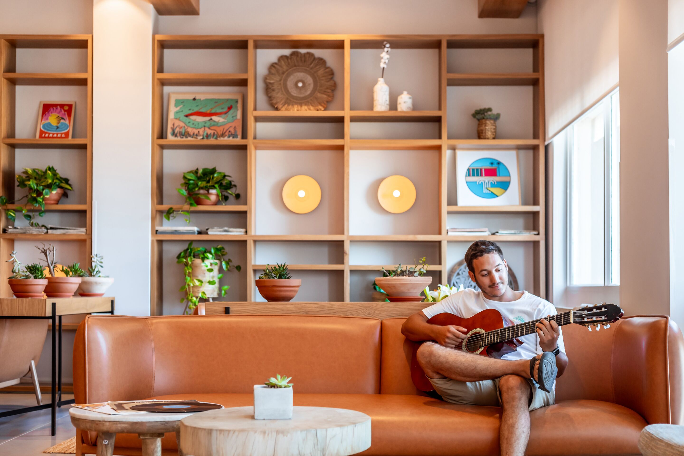 A person sitting on a leather couch playing a guitar in a room with wooden shelves containing plants and decorative items.