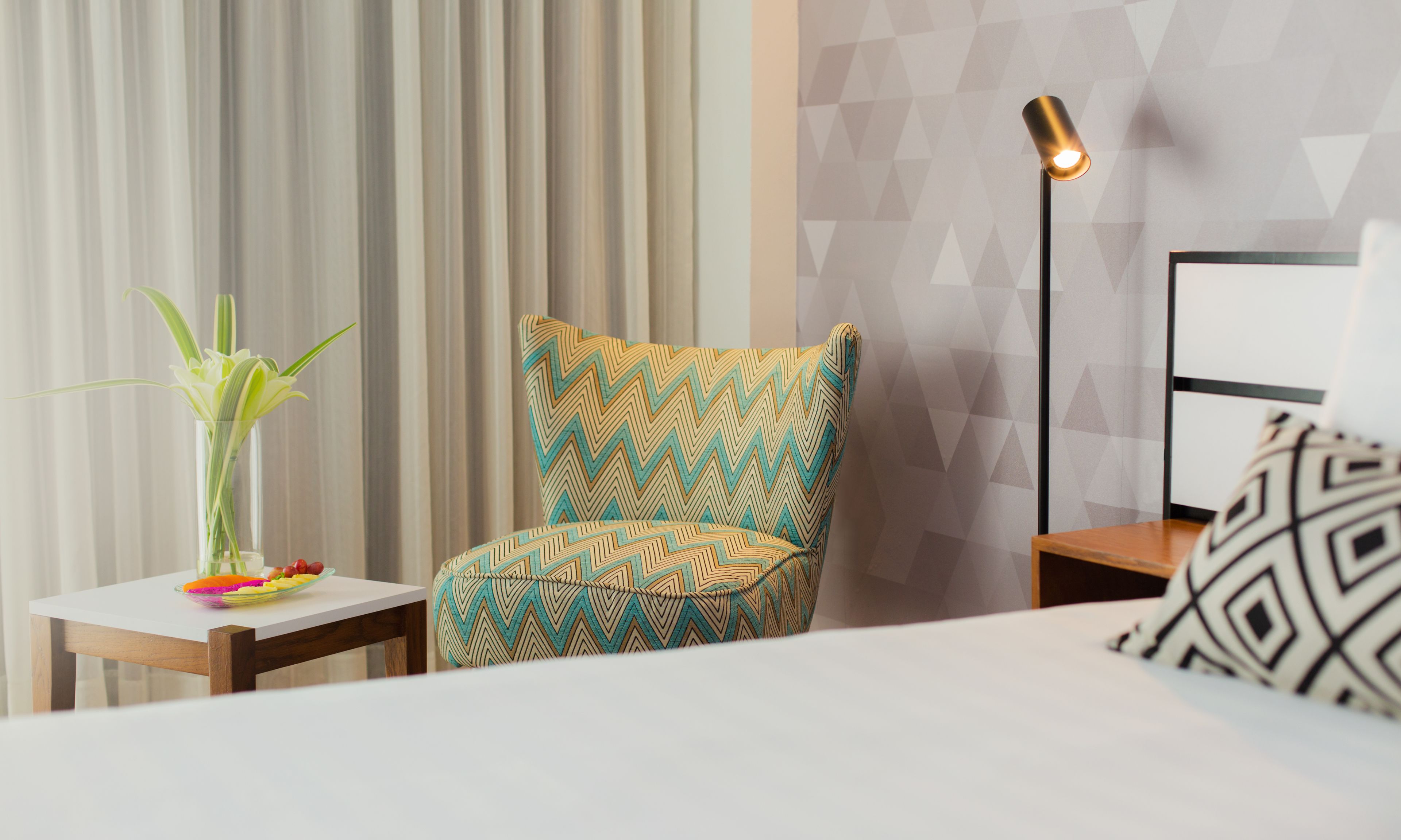Habitación de hotel con una cama, un sillón estampado, una mesita con un jarrón de flores, una lámpara y una pared con diseño geométrico.