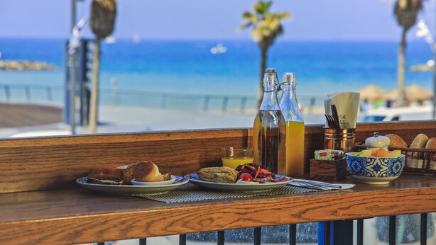 Una mesa de desayuno con vista al océano, que incluye pan, un tazón de fruta y una botella de jugo.